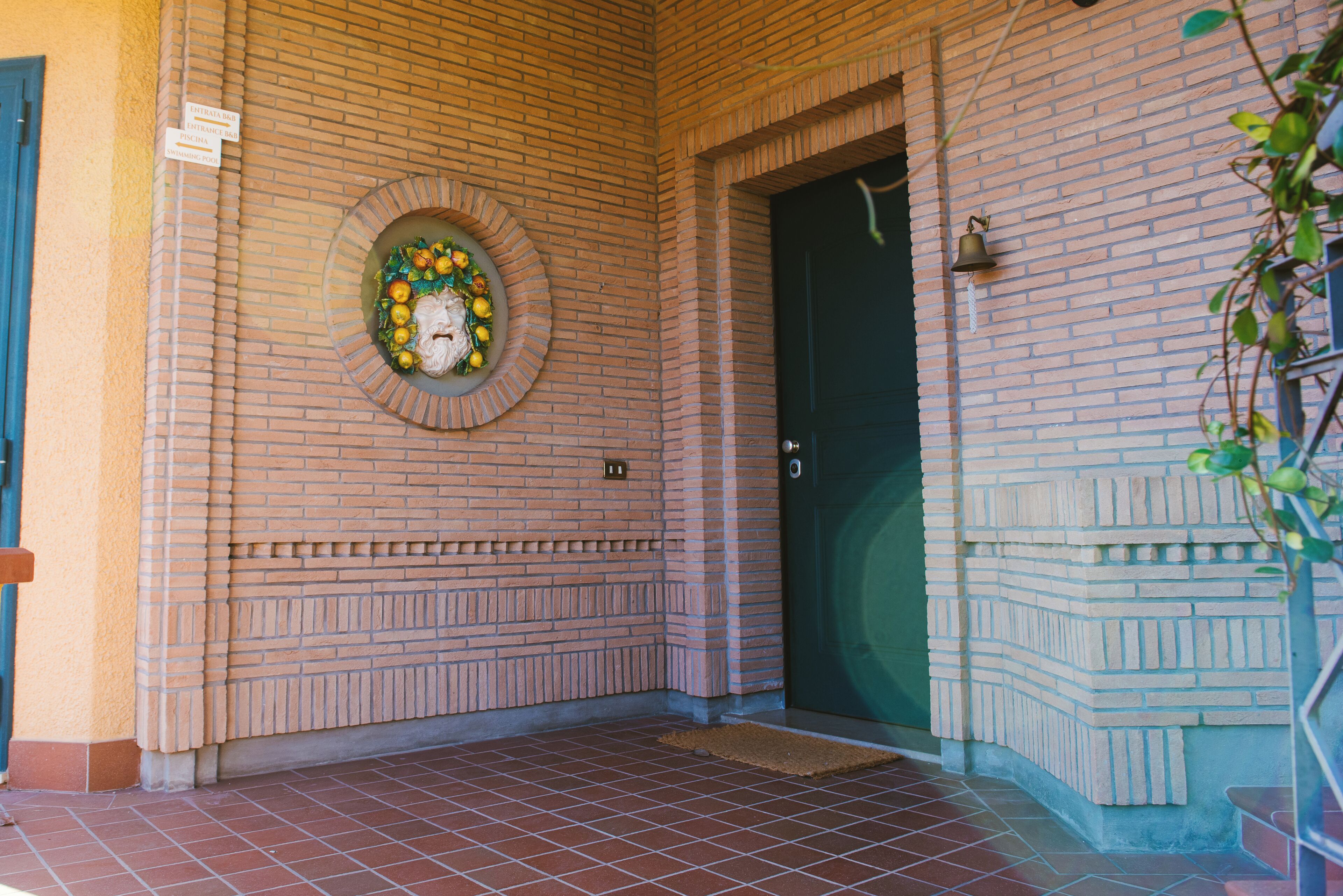 Entrada de un edificio de ladrillo con un elemento decorativo circular en la pared, una puerta verde y una pequeña campana encima.