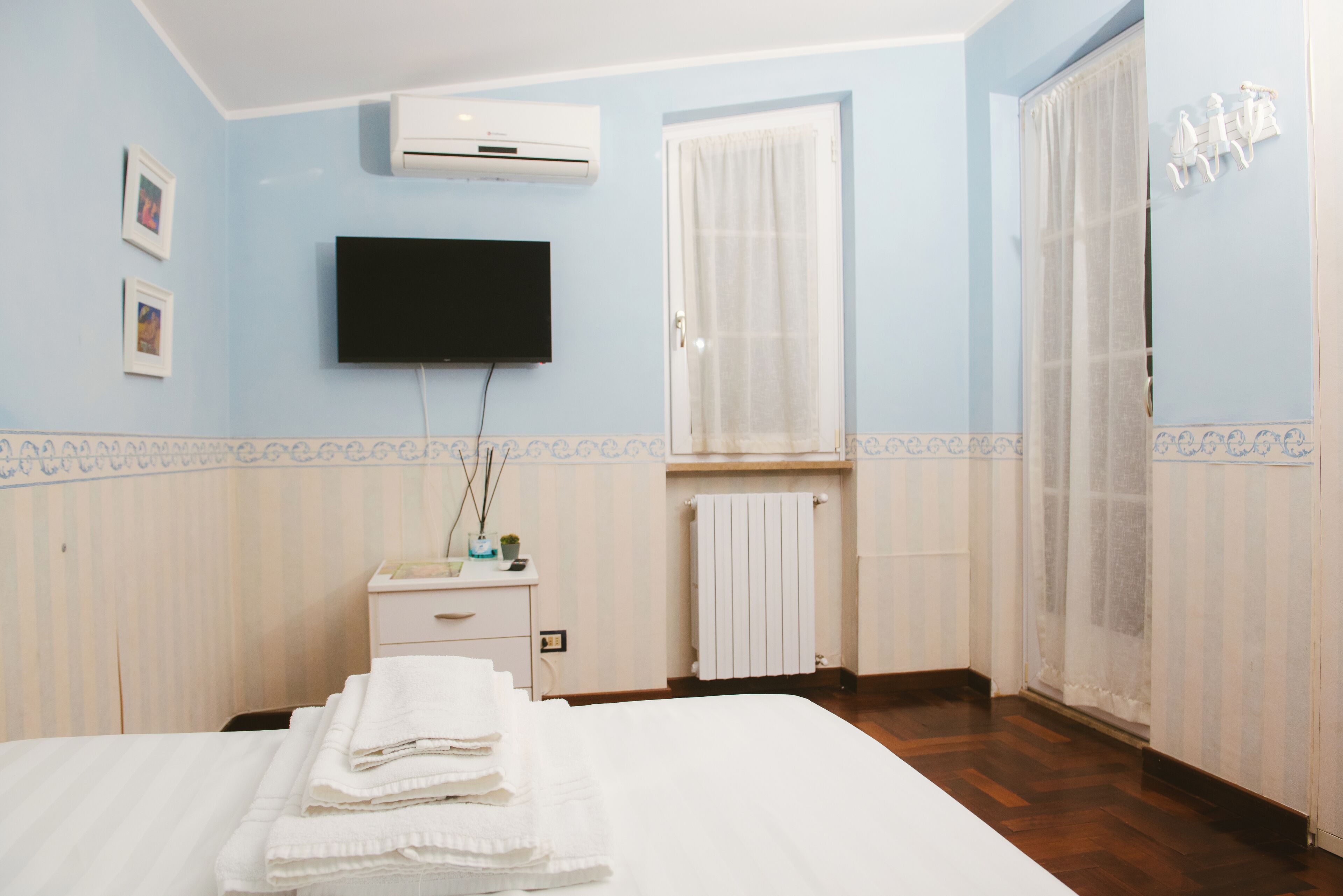 A hotel room with a bed, a TV, a window with curtains, a radiator, and a small table with a plant.