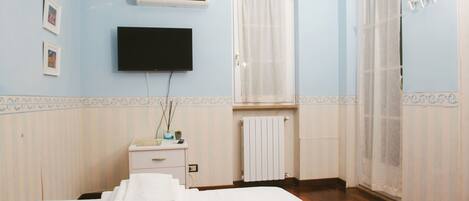 A hotel room with a bed, a TV, a window with curtains, a radiator, and a small table with a plant.