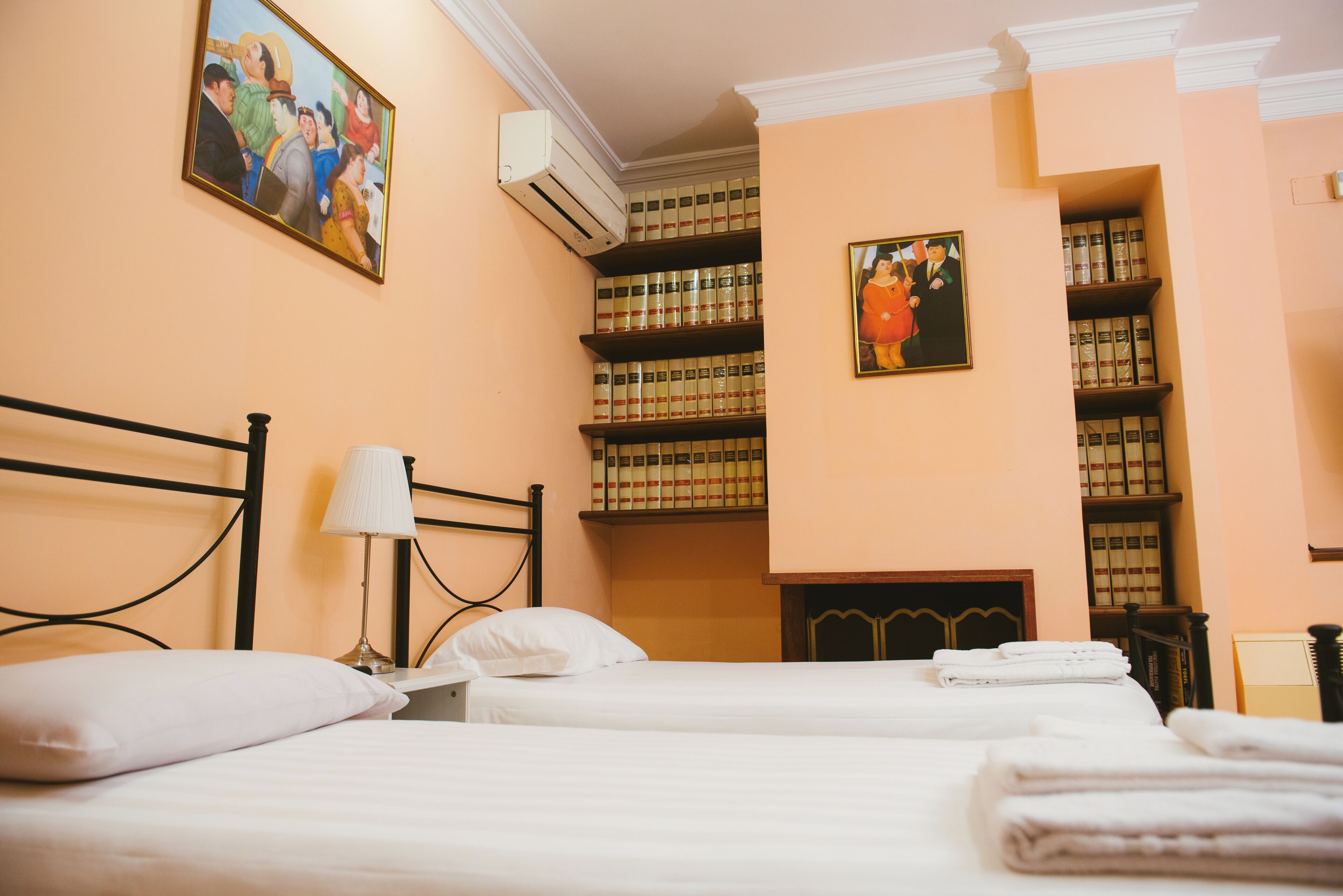 A hotel room with two beds, a bedside lamp, a wall-mounted air conditioner, and shelves with books.
