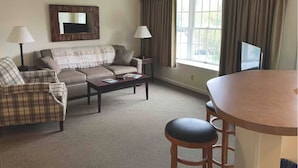 Een woonkamer met een bank, fauteuil, salontafel en een eettafel met stoelen. Er is een televisie op een dressoir en een raam met gordijnen.