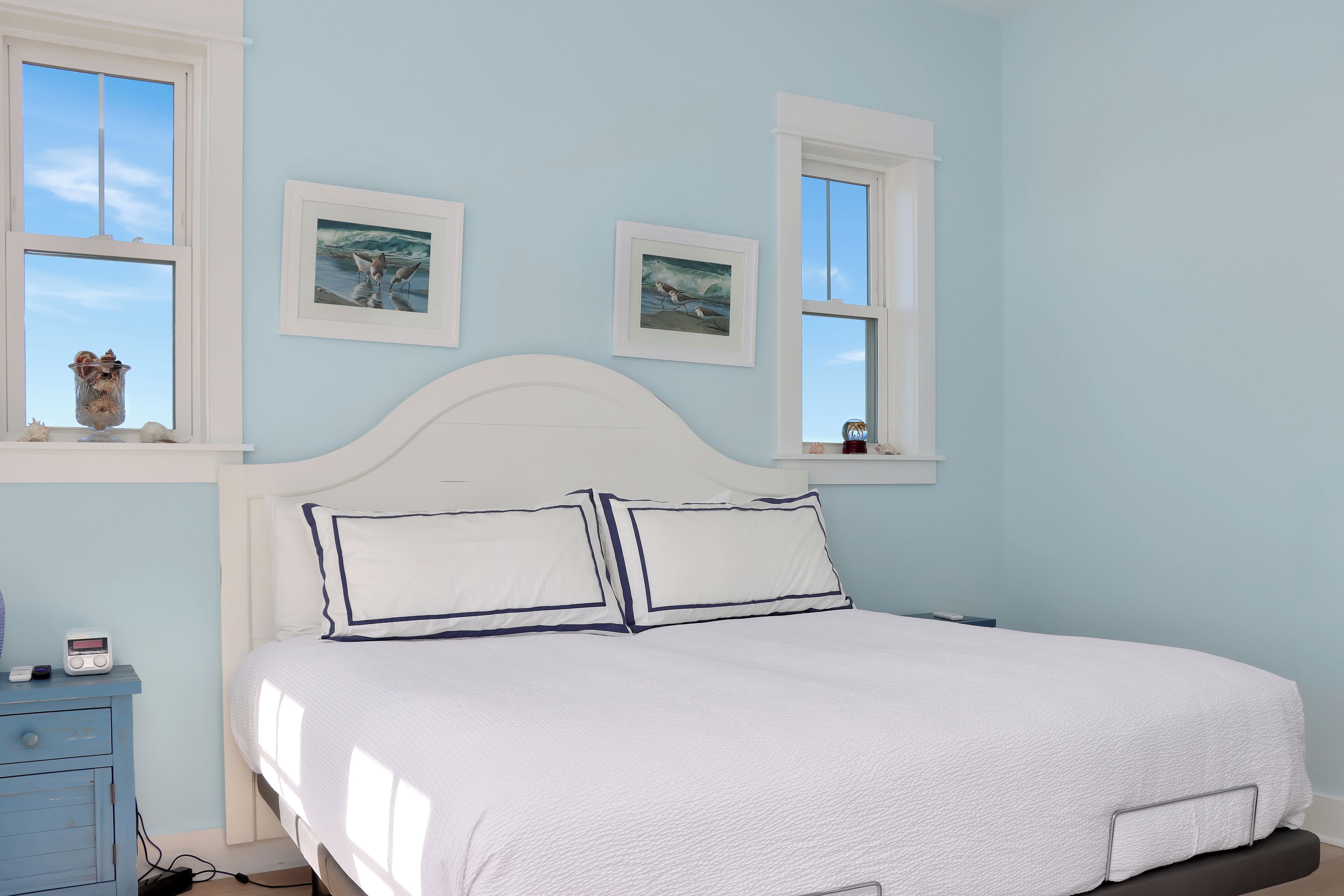 Une chambre à coucher avec un lit blanc, deux tableaux encadrés au mur et une fenêtre donnant sur le ciel.