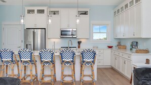 A modern kitchen with white cabinets, a stainless steel refrigerator, a microwave, and a large window. There are four wicker bar stools with blue and white patterns. - 🌊Dog Friendly Home with Private Pool-Steps to the Beach-Salty Owl🌊 (Cape San Blas)