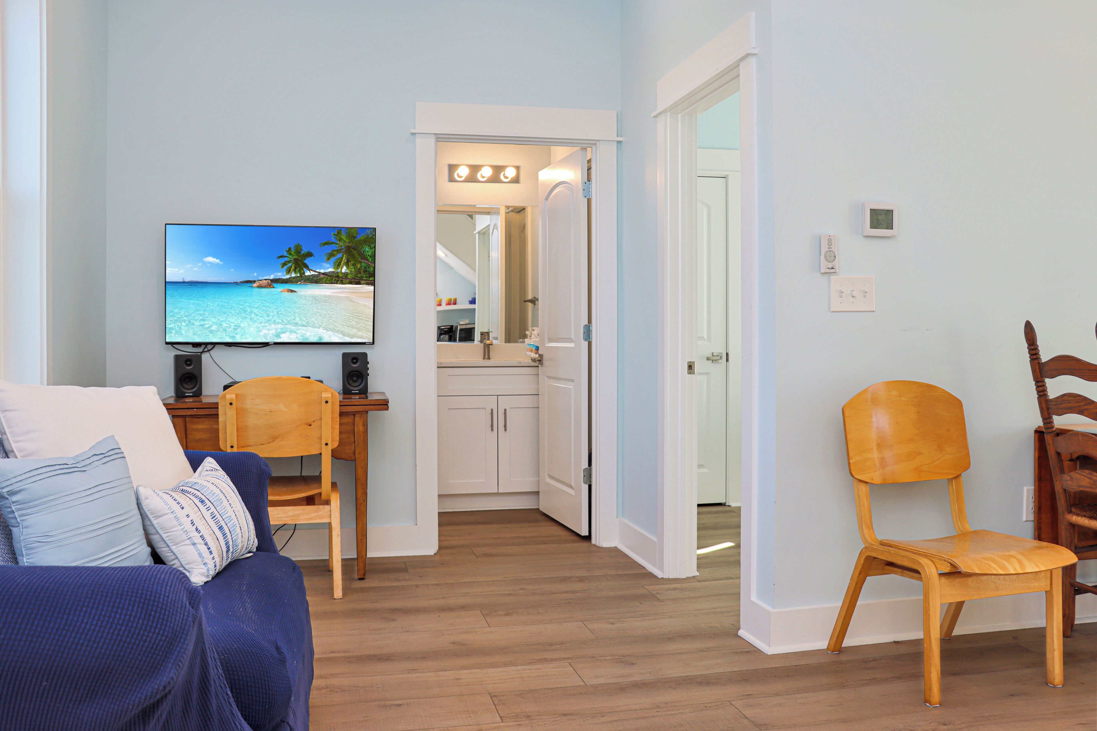 Un salón con una televisión mostrando una escena de playa, una silla de madera, un sofá con cojines estampados y una cocina visible a través de una puerta abierta.