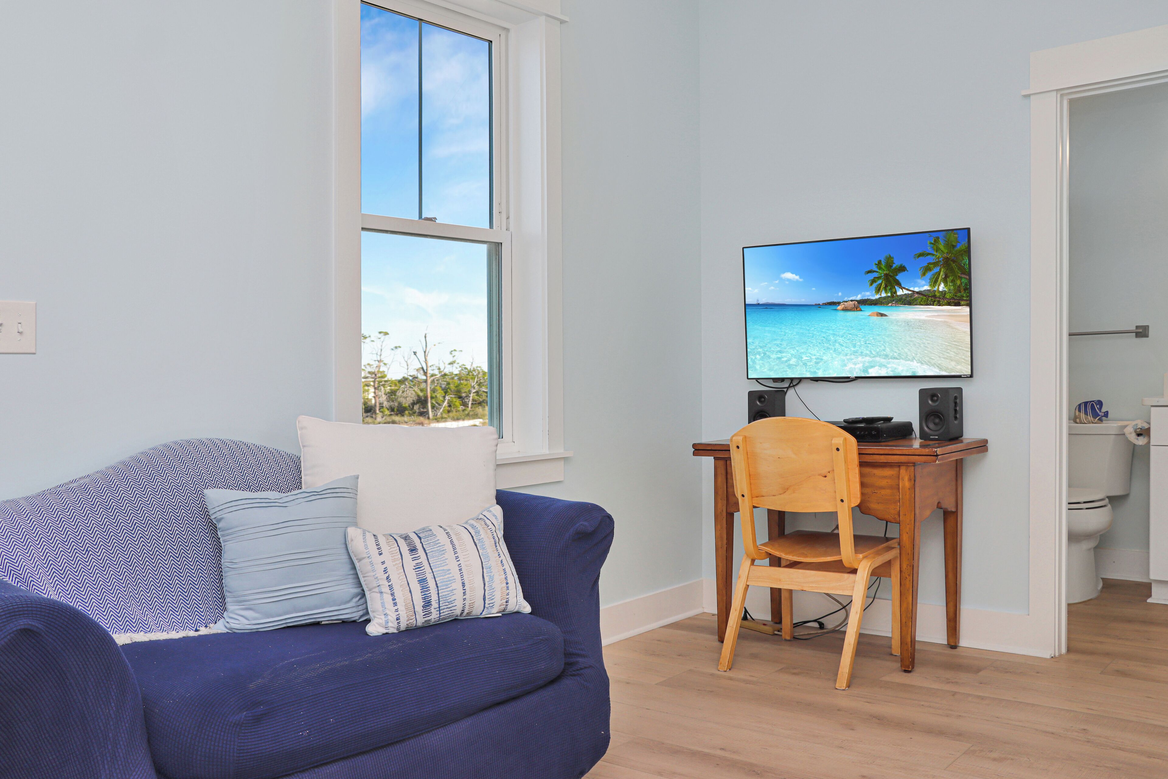 A living room with a blue sofa, a wooden chair, a television displaying a beach scene, and a window overlooking a view.