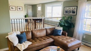 A living room with a brown leather sectional sofa, a wooden coffee table, and a large potted plant. - Spacious Charming Kennebunk Home (Kennebunk)