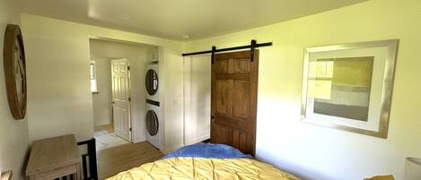 Een slaapkamer met een bed, een houten deur, een wasmachine en een klein tafeltje.