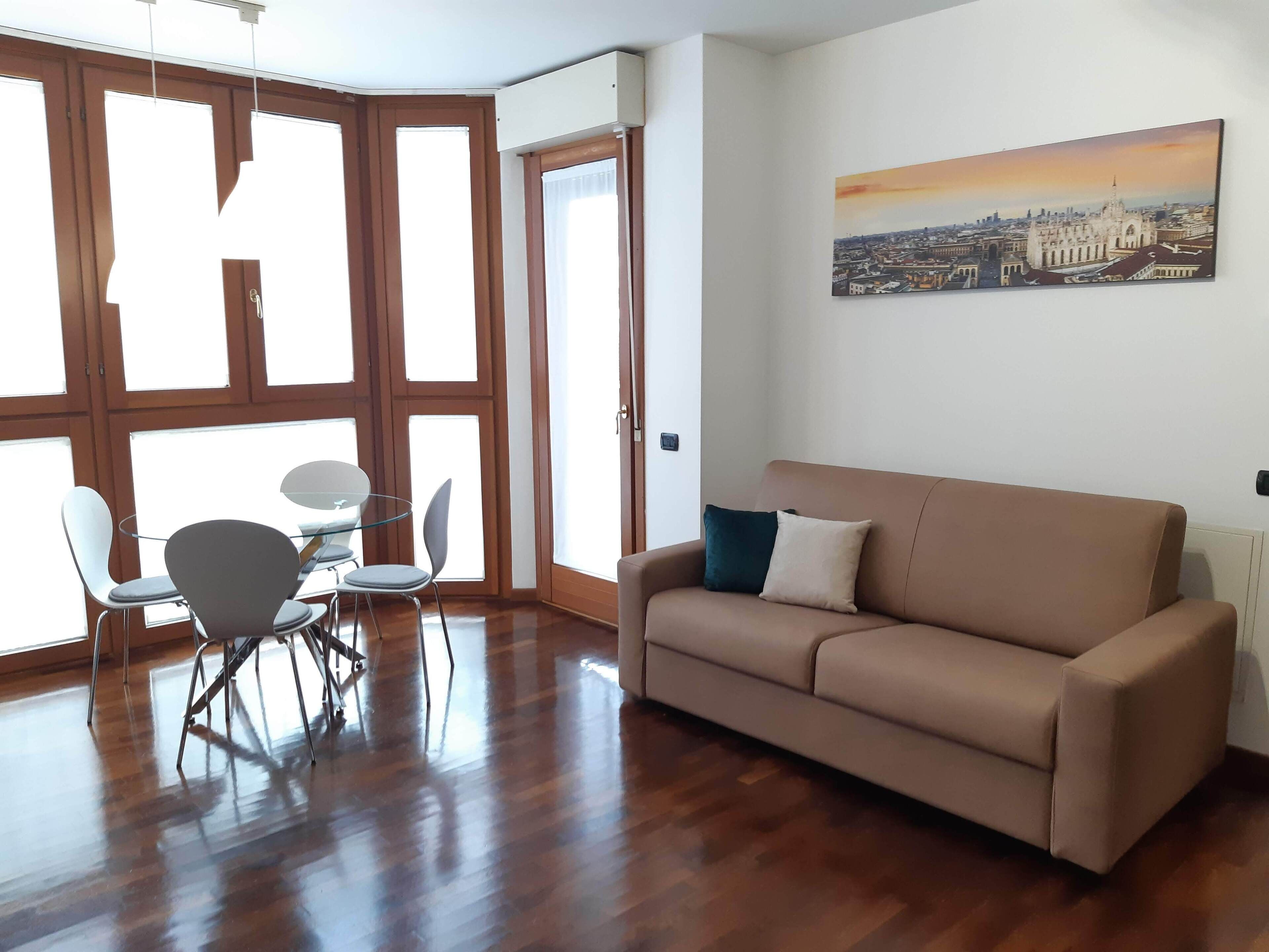 Una habitación con piso de madera, un sofá color beige, una mesa de vidrio con sillas y un ventanal con vistas a la ciudad.