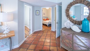 A hallway with a tiled floor, a bed with orange pillows, a mirror with a rustic frame, and a wooden console table with decorative items.