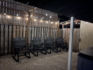 Relax in comfort with four Adirondack chairs arranged on a gravel surface, surrounded by a wooden fence and ambient lighting.