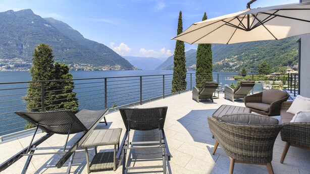 A terrace with wicker furniture, a table, and an umbrella overlooking a lake with mountains in the background.