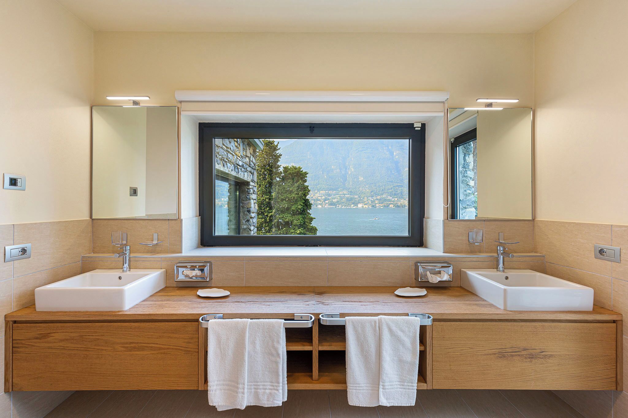 A modern bathroom with two sinks, a large window with a view of mountains, and wooden cabinetry.
