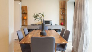 A dining area with a wooden table, six chairs, a vase with a plant, a microwave, and shelves with glassware and a red mug.