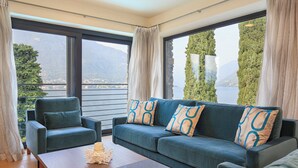 A modern living room with a sofa and armchair, large windows with curtains, and a view of a lake and mountains.