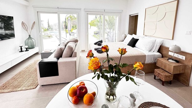 Une chambre d’hôtel moderne avec un grand lit, un canapé, une table à manger agrémentée de fruits et de fleurs, et une vue sur l’extérieur.