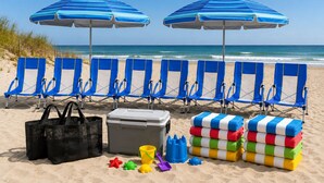 Ein Strand mit zwei blauen Sonnenschirmen, einer Reihe blauer Liegestühle, einer schwarzen Tasche, einer grauen Kühlbox und bunten Strandspielzeugen.