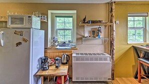 A compact kitchen with a refrigerator, microwave, and a portable air conditioner. There are windows providing natural light, and a small table with kitchenware. - Duck Lake Cabin Rental Near GNP (Babb)