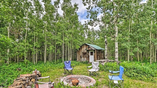 A rustic cabin surrounded by a dense forest, with a fire pit and several chairs in the foreground. - Duck Lake Cabin Rental Near GNP (Babb)