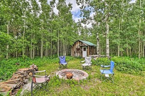 Une cabane rustique entourée d’une forêt dense, avec un foyer et plusieurs chaises au premier plan.