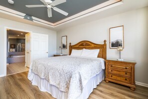 Une chambre à coucher avec un grand lit, des tables de chevet en bois, un ventilateur de plafond et une porte donnant sur une salle de bain.