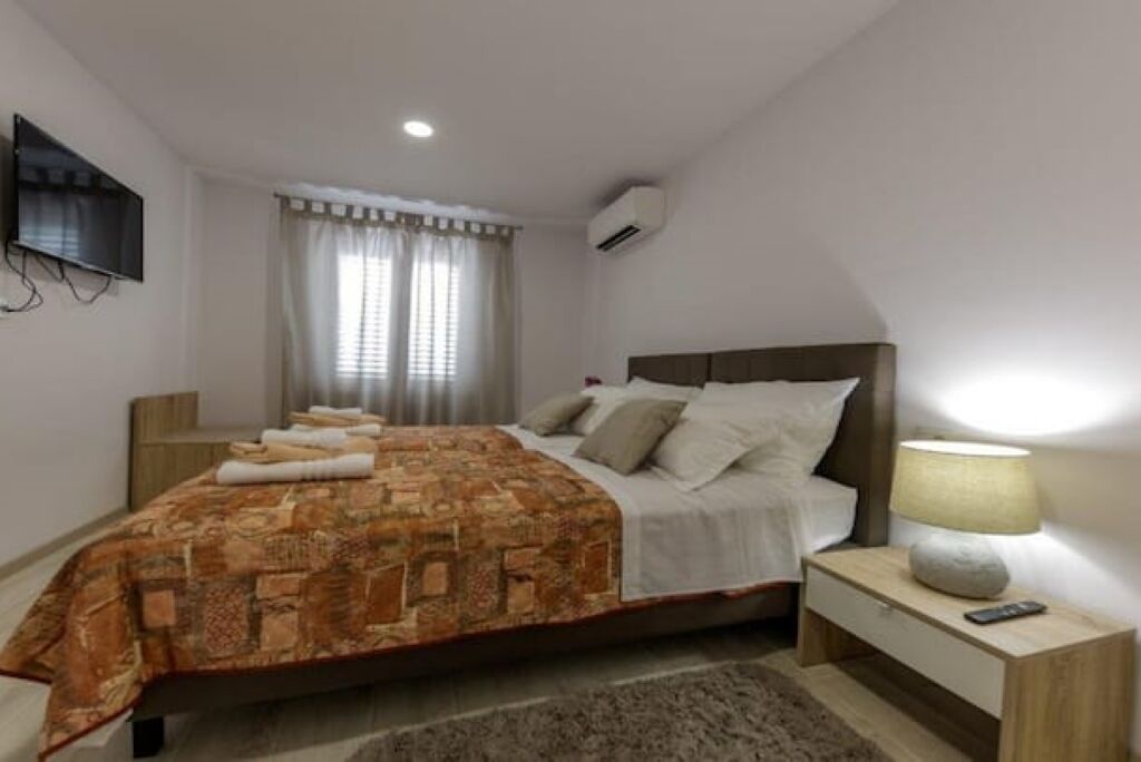 A bedroom with a bed, a nightstand, a lamp, a TV, and a window with curtains.