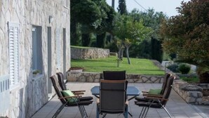 Une terrasse extérieure avec un mur en pierre, une table et des chaises, et une vue sur un jardin aménagé.