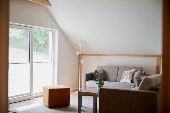 A modern living room with a grey sofa, a small wooden table, and a bench. There is a large window providing natural light and a view of greenery outside. - GUTSHOF Schirgiswalde (Schirgiswalde-Kirschau)