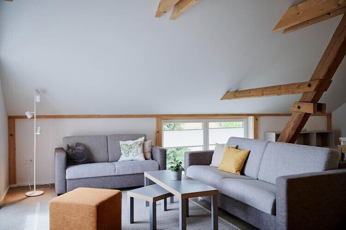 Een moderne loft-woonkamer met schuine plafonds, ingebouwde houten balken, twee grijze banken met kussens, een kleine salontafel en een vloerlamp.