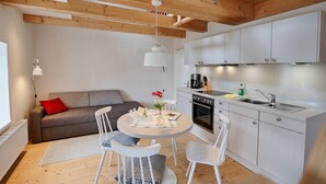 Una cocina compacta con armarios blancos, una zona de comedor con una mesa redonda y sillas, y un sofá gris con un cojín rojo.
