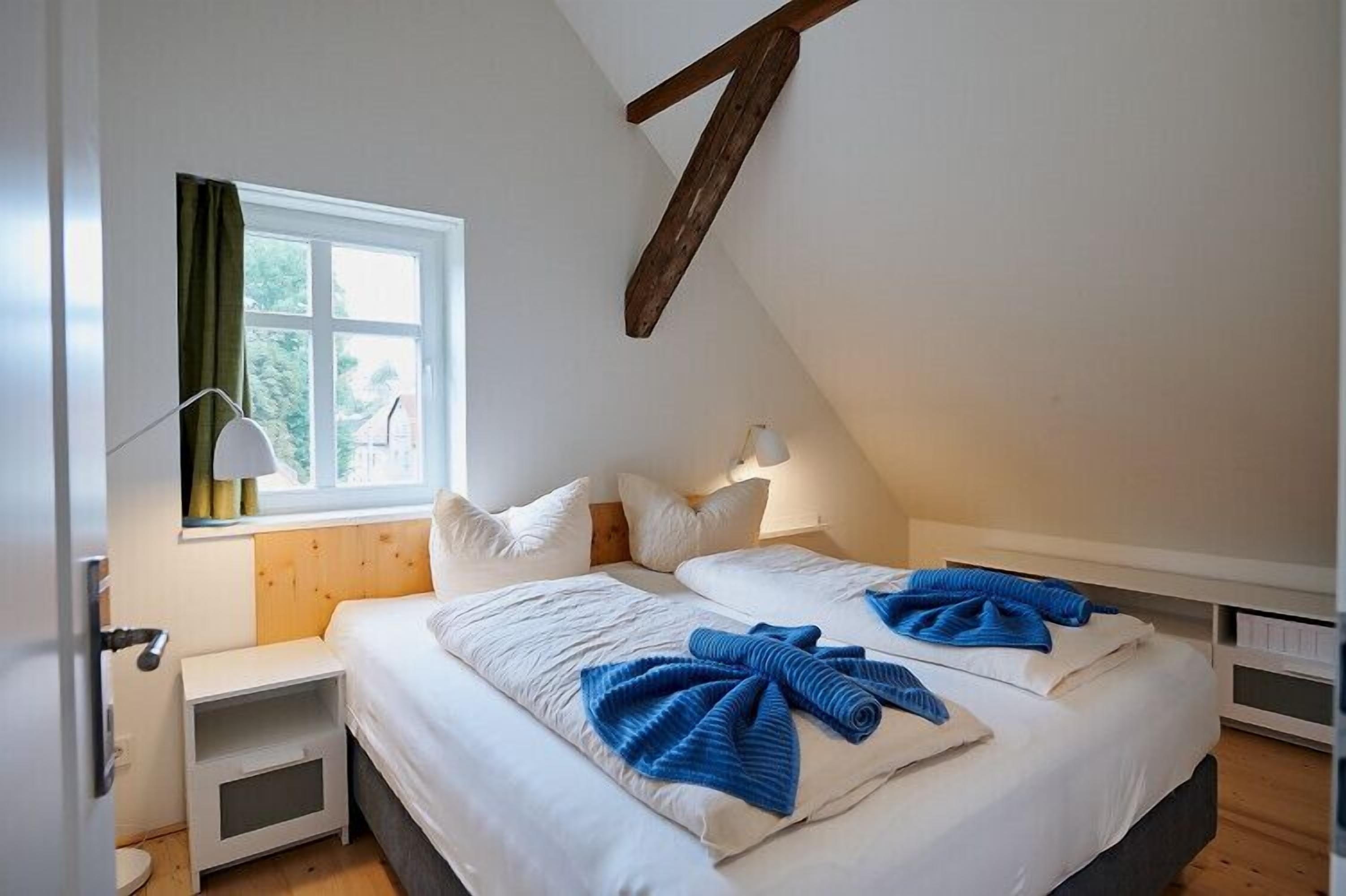 Une chambre avec un plafond en pente, une fenêtre avec des rideaux, une table de chevet avec une lampe, et un lit avec une literie blanche et des serviettes bleues.