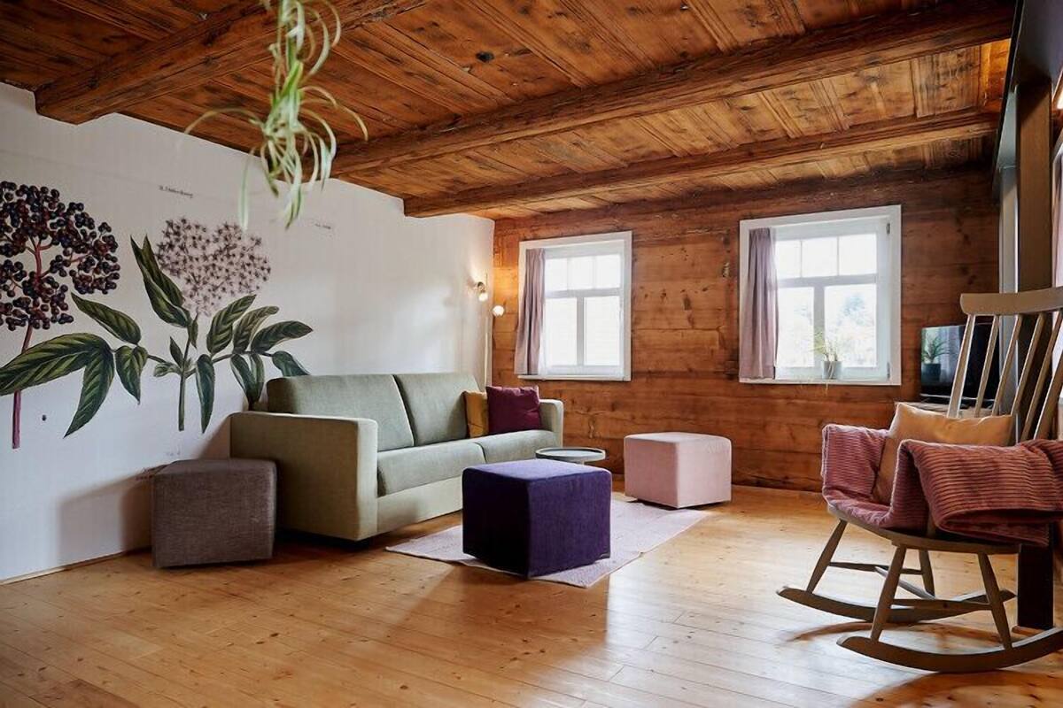 Een gezellige woonkamer met een houten plafond, een groene bank, een schommelstoel en een wand met botanische illustraties.
