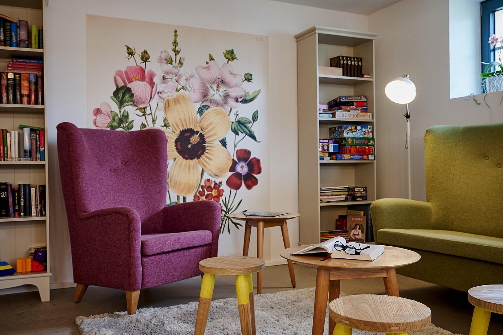 Un salón acogedor con un amplio mural floral, un sillón morado, un sofá verde y una estantería llena de libros y juegos.