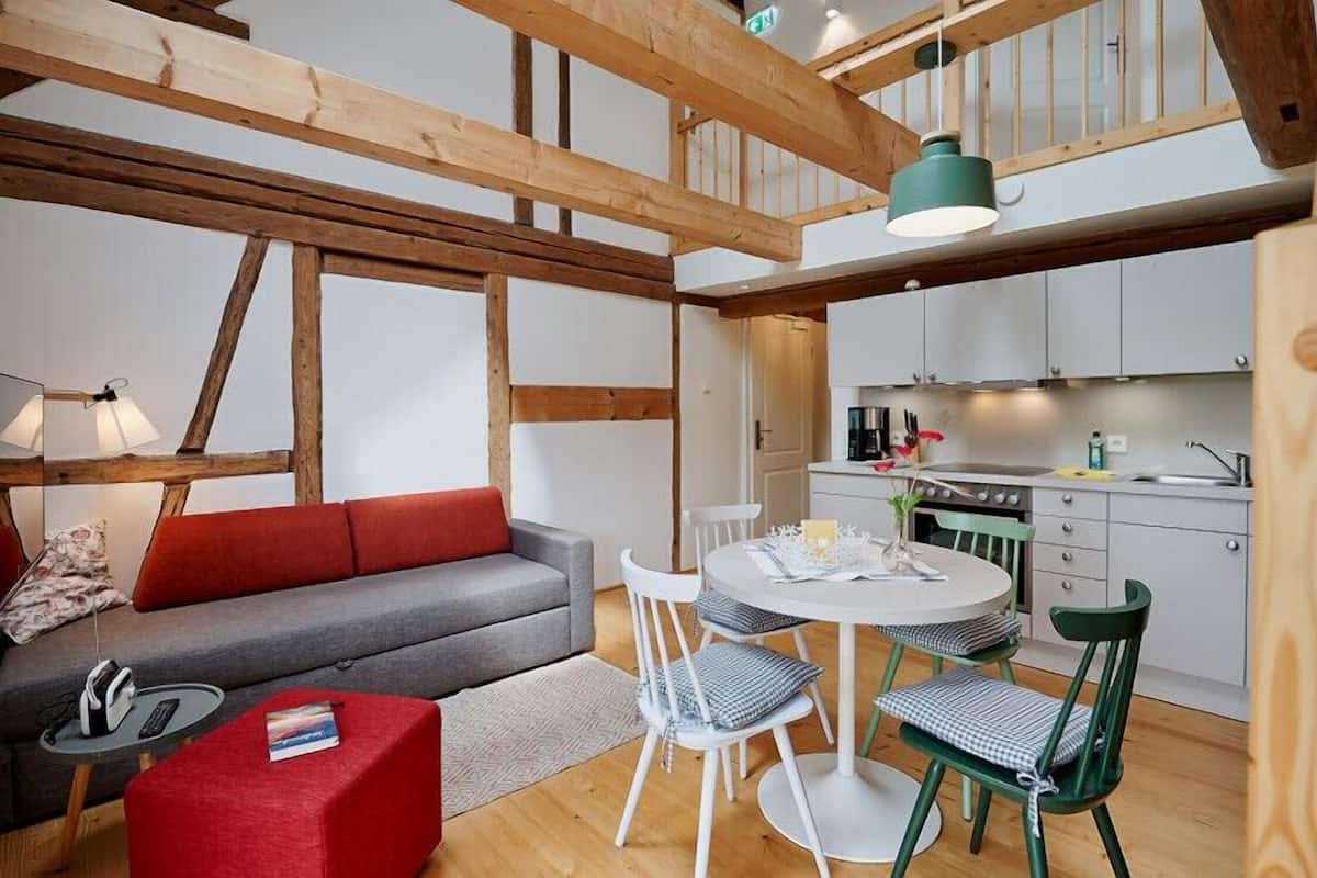 Een gezellige zithoek met een bank, een klein rond tafeltje en stoelen. In de achtergrond is een keuken met kastjes en een fornuis te zien.