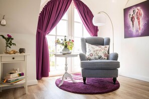 A room with a grey armchair, a small round table with a vase of flowers, a bookshelf, and a window with purple curtains.