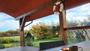 A wooden deck with a table and chairs, a lifebuoy, and a view of a grassy area with trees. - Comfortable holiday home, private garden, sauna (Smołdzino)