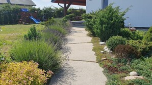 A pathway leading to a building with a covered entrance, surrounded by various shrubs and plants. - Comfortable holiday home, private garden, sauna (Smołdzino)