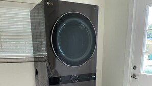 Lavadora y secadora LG compacta de dos puertas, con carga frontal, ubicada en una habitación con puerta blanca y ventana con persianas.