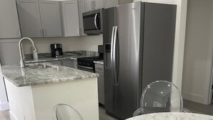 Una cocina moderna con gabinetes grises, electrodomésticos de acero inoxidable, encimera de mármol y sillas de plástico transparente.
