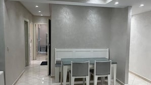Een eetgedeelte met een glazen tafel en witte stoelen, een ingelegd plafond en een gang naar een andere kamer.