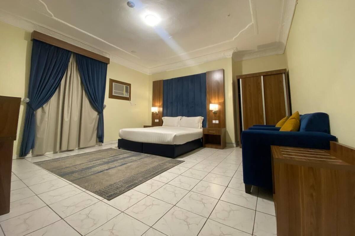 A hotel room with a bed, a sofa, a wooden side table, and a blue curtain.