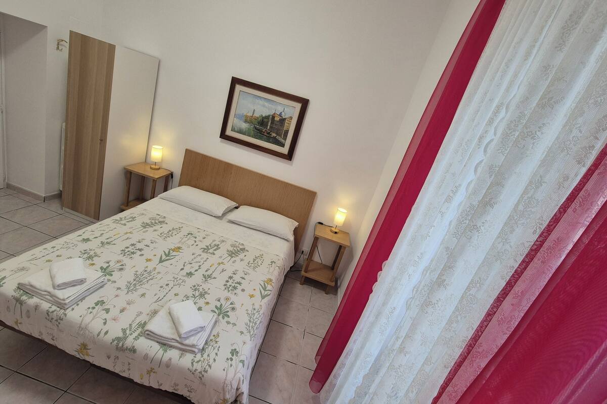 A hotel room with a bed, two bedside tables, a nightstand, a painting on the wall, and a red curtain.