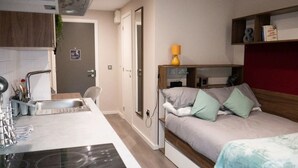 Une chambre d’hôtel compacte comprenant un coin cuisine, un lit avec des oreillers, une table de chevet et une étagère avec des livres.