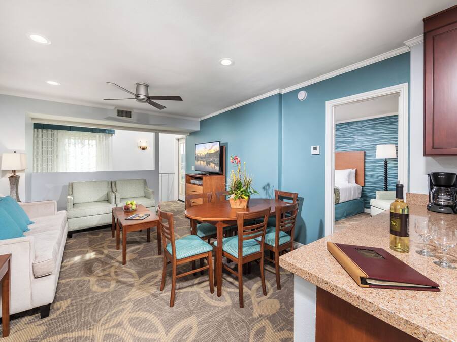 A modern living room with a sofa, coffee table, and dining area with chairs. There is a ceiling fan, a TV, and a kitchen area with a coffee maker and wine glass.