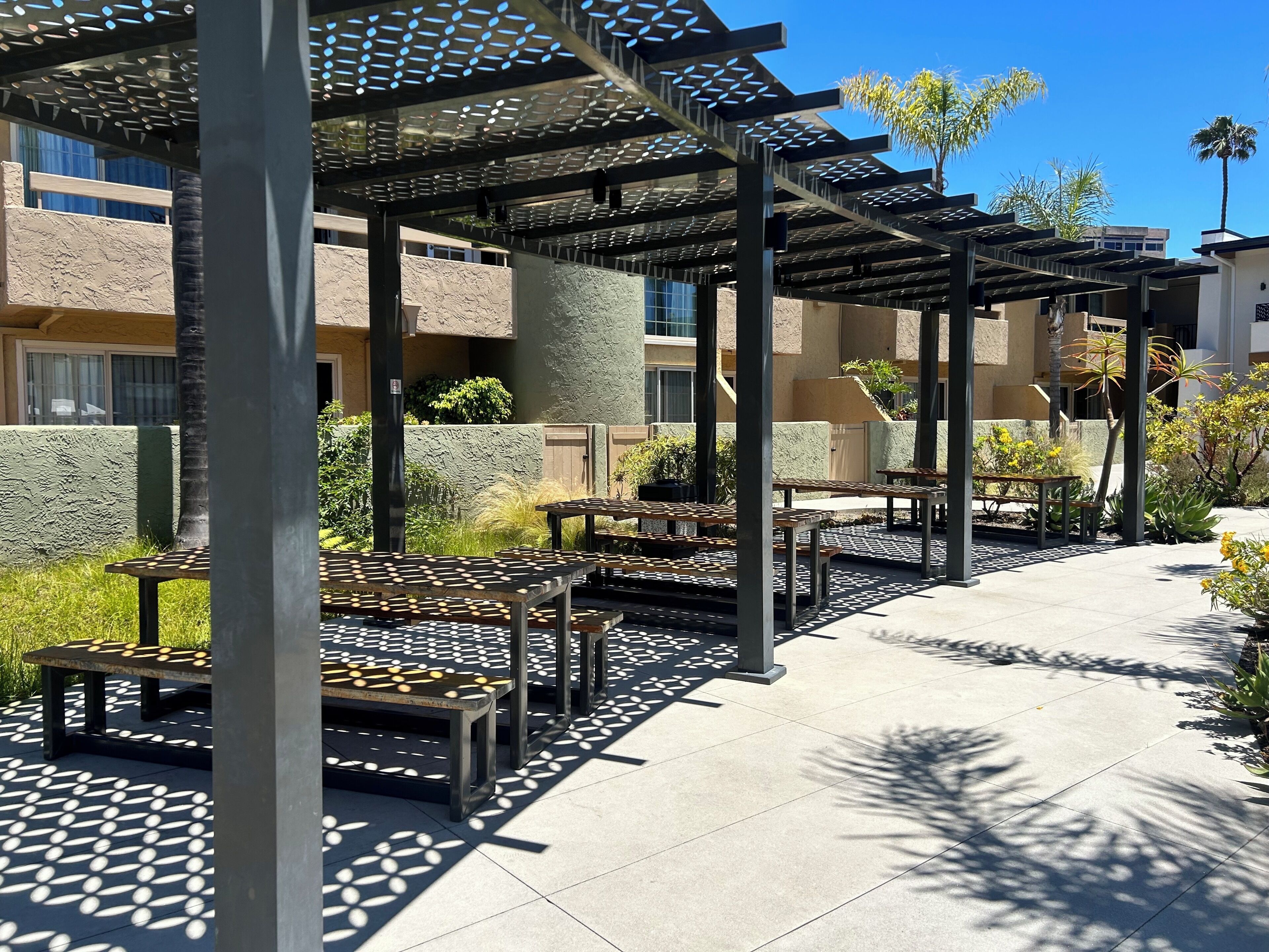 Un área de patio al aire libre con mesas y bancos, una estructura de pérgola y vistas a un edificio al fondo.