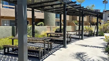 Un área de patio al aire libre con mesas y bancos, una estructura de pérgola y vistas a un edificio al fondo.