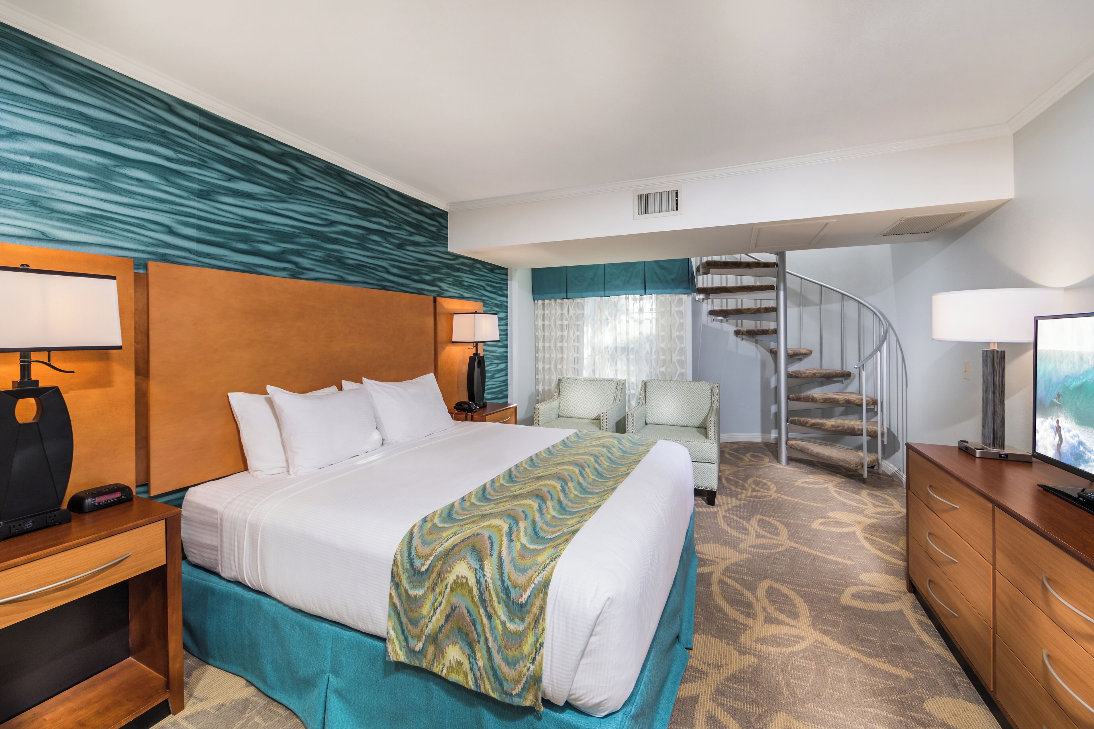 A hotel room with a large bed, a wooden headboard, a desk with a lamp, a chair, a TV, and a spiral staircase.