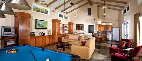 A spacious living area with a pool table, a sofa, and a red armchair. There is a kitchen in the background with a dining table and chairs.