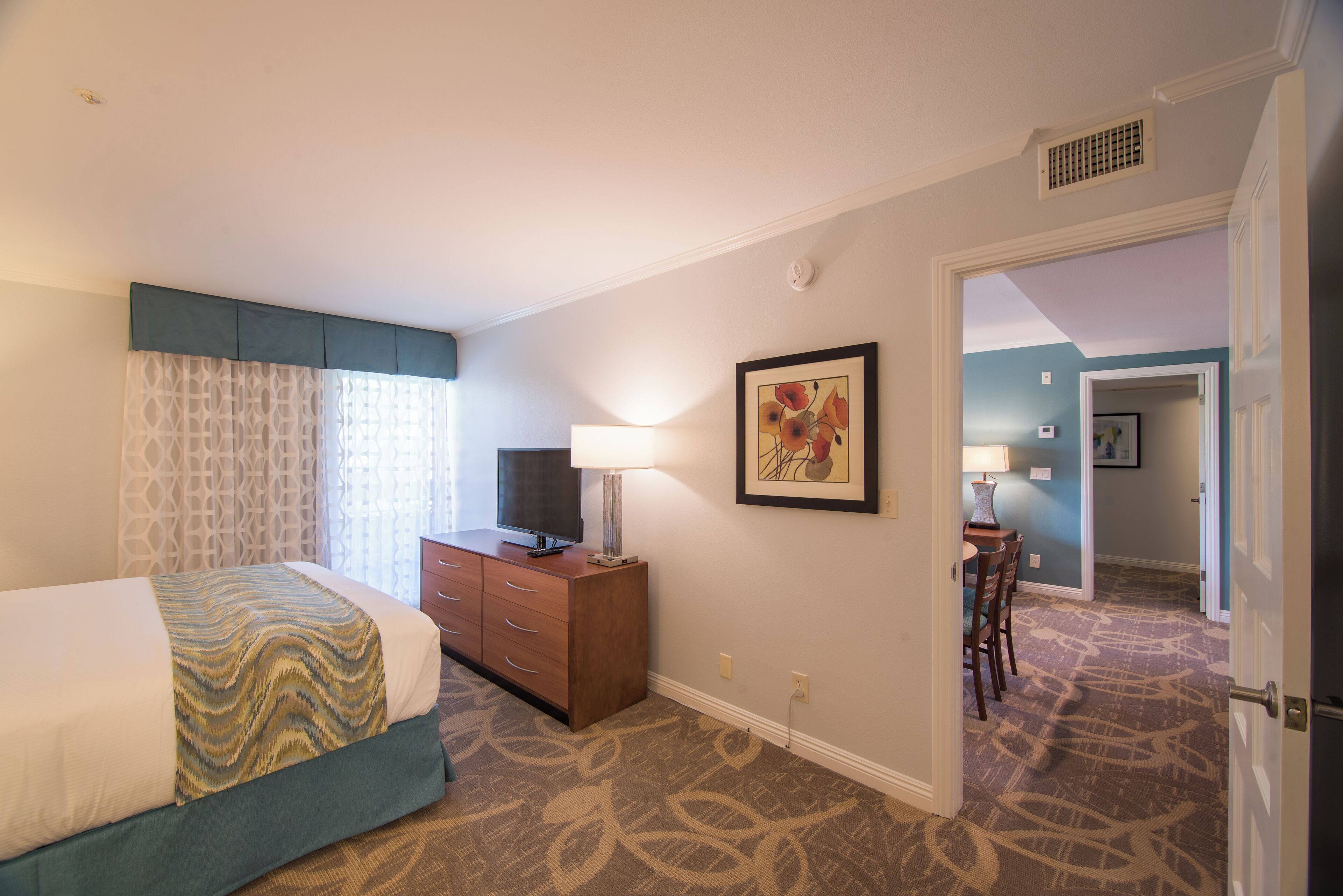 A hotel room with a bed, a desk, a chair, a television, a lamp, and a painting on the wall.