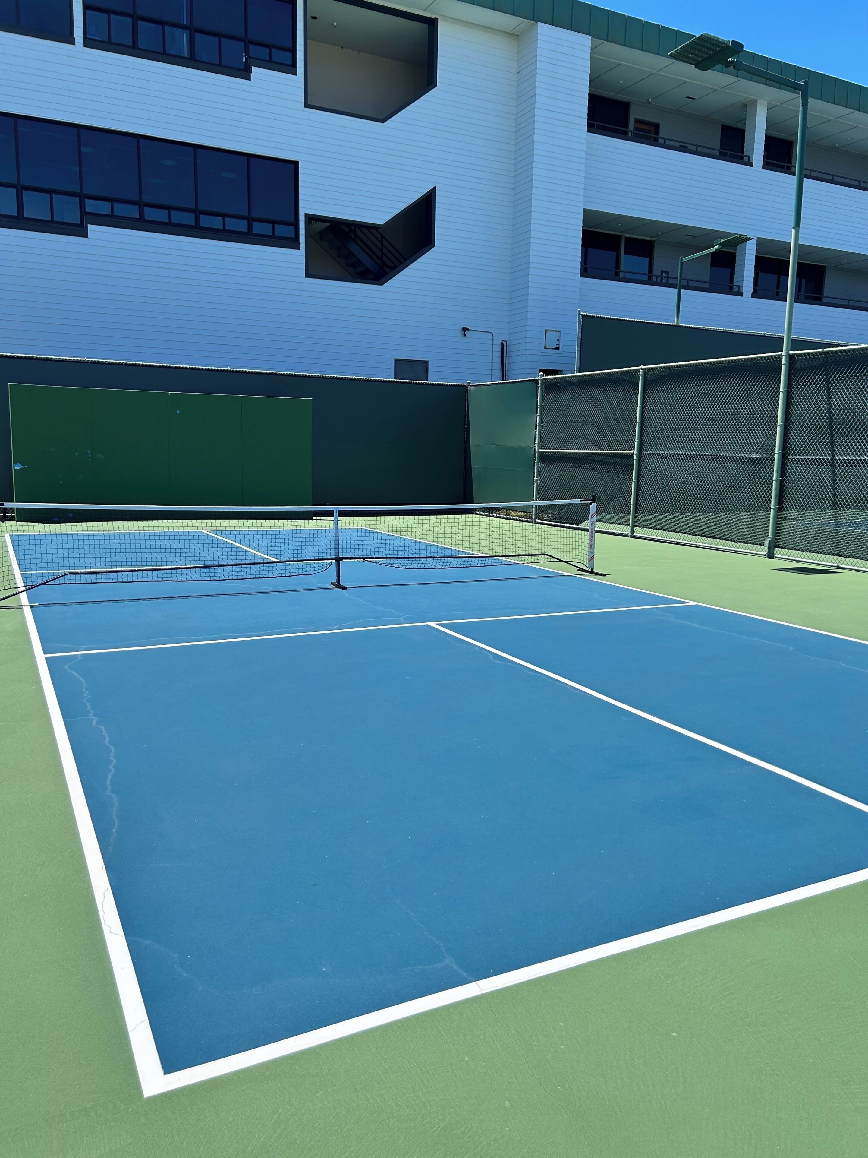 Una cancha de tenis al aire libre con red, rodeada por una cerca verde, y contigua a un edificio blanco con numerosas ventanas.
