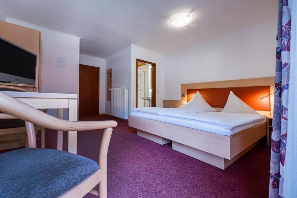 A hotel room with a bed, a desk with a chair, a TV, and a bathroom visible through an open door.
